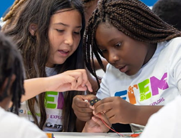 Two girls wearing STEM t-shirts work together on a small electronics project at a table, focusing closely on the components in their hands.